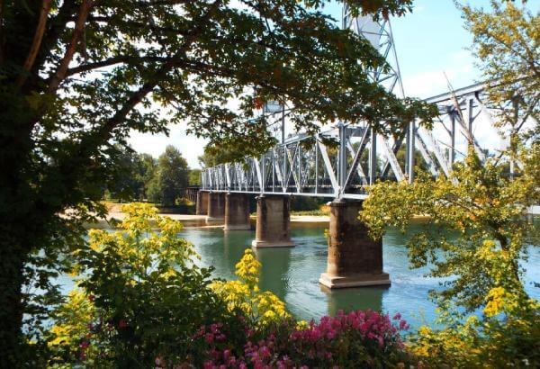 A metal bridge spans across a river with surrounding greenery and blooming flowers in the foreground. Salem, Oregon