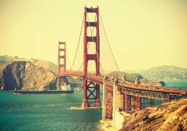 A large red suspension bridge spans a body of water, with hills in the background under a clear sky. A ship is passing under the bridge. A large red suspension bridge spans a body of water, with hills in the background under a clear sky. A ship is passing under the bridge.