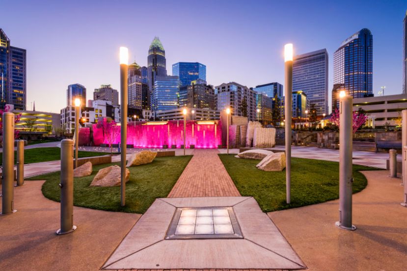 Urban park scene at dusk with a pink-lit water feature, surrounded by modern buildings and illuminated lamp posts, set under a clear sky.