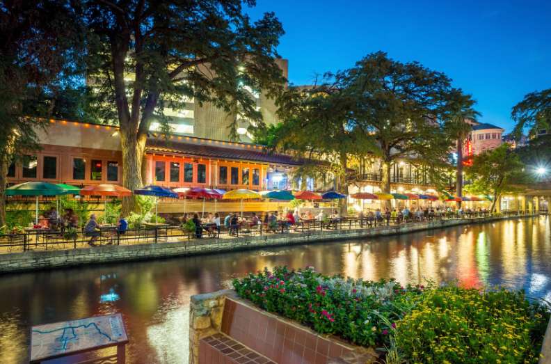 People dine under colorful umbrellas along a tranquil riverside lined with trees and buildings at dusk.