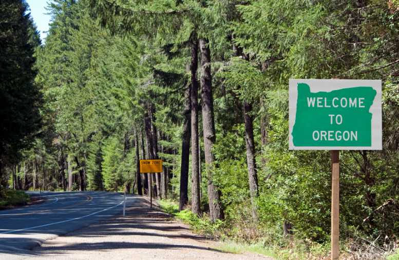 Welcome to Oregon" sign beside a winding road lined with lush green trees. Welcome to Oregon" sign beside a winding road lined with lush green trees.